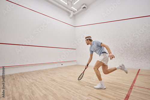 athletic and redhead sportsman with racquet playing squash inside of court, motion shot