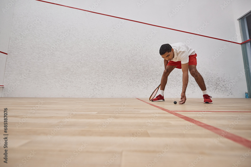 african american man in red shorts holding racquet and picking up squash ball inside of court