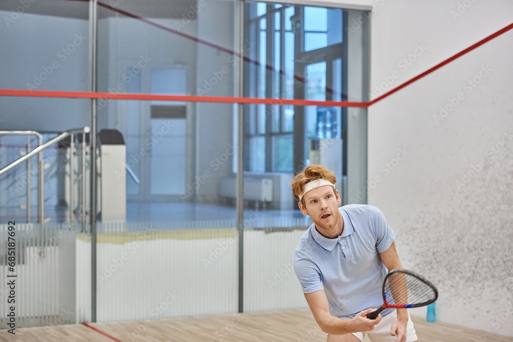 energetic redhead sportsman holding racquet while playing squash inside of court, sport challenge