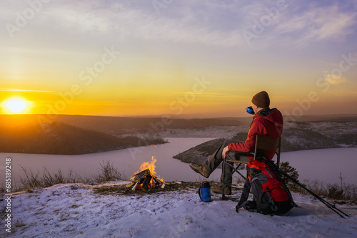 man hiker sitting near bonfire and drinking tea during camping in winter at sunset.