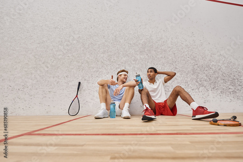 two interracial sportsmen with squash racquets and bottles with water sitting on floor inside court