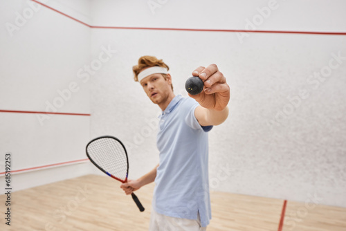 redhead man in sportswear showing squash ball and holding racquet while standing inside of court © LIGHTFIELD STUDIOS
