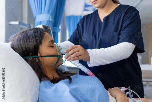 Biracial female doctor applying oxygen mask to female patient in bed in hospital room photo