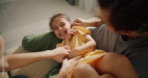 Close up shot of a happy little girl in a yellow dress laughing and rejoicing while being tickled by her parents sitting on a light brown in a modern apartment
