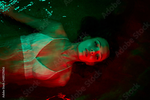 Woman floats in lagoon  red illumination surrounds her  night setting conveys solitude  introspection  eerie atmosphere  concept of inner world exploration and duality of self.