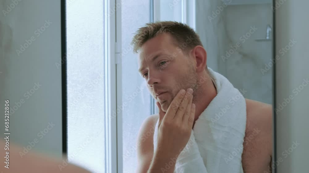 Young handsome young man toching his bristle on the face standing in the bathroom with a towel and looking at mirror. Preparation for daily morning routine male hygienic beauty procedure Young handsome young man toching his bristle on the face standing in the bathroom with a towel and looking at mirror. Preparation for daily morning routine male hygienic beauty procedure