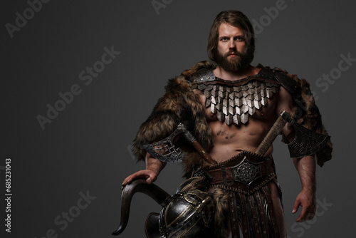 A fierce bearded Viking warrior in fur and light armor, with a helmet attached to his belt, holding a large two-handed axe on a gray background © Fxquadro