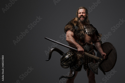 A fearless Viking warrior with a majestic beard adorned in fur and light armor, carrying an axe and shield, standing against a gray background