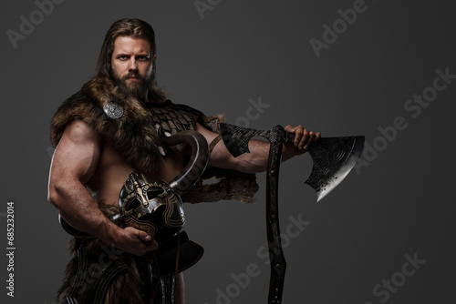 A fierce bearded Viking warrior in fur and light armor, with a helmet attached to his belt, holding a large two-handed axe on a gray background