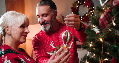Close up of joyful senior Caucasian couple husband giving xmas gift box to beloved wife at home and decorating tree with christmas toy preparing for New Year celebration. Merry Christmas concept