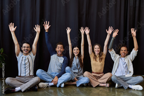 excited stylish multiethnic team sitting and waving hands near black drape in office, group photo