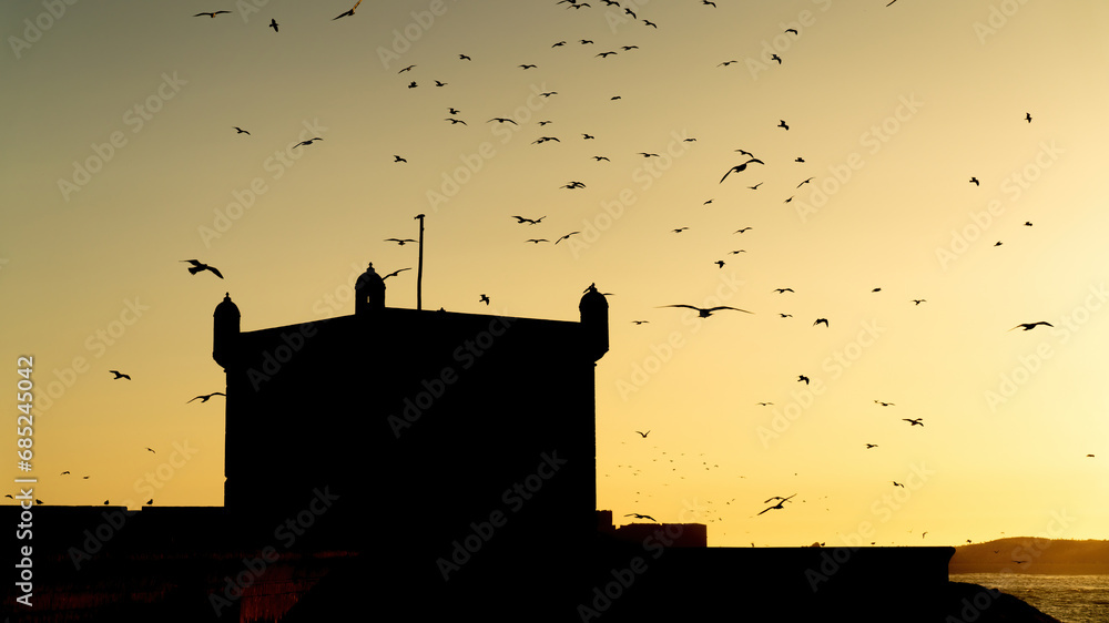 Silhouette of Sqala du Port at sunset, a historic fortress overlooking the Atlantic Ocean at the fishing port of Essaouira with many seagulls flying overhead, Morocco.