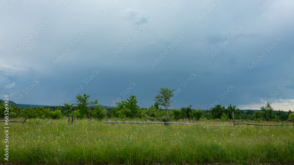 A grassy field with an old wooden fence in a rural area. Behind them lies an oak forest. Overcast weather. Storm is approaching. Romania.
A grassy field with an old wooden fence in a rural area. Behind them lies an oak forest. Overcast weather. Storm is approaching. Romania.