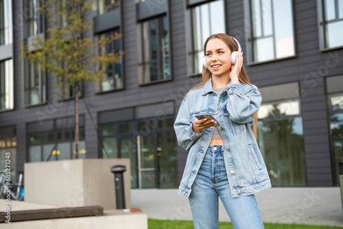 A cheerful young lady, sporting headphones and clutching a mobile device, beams with delight in the street 