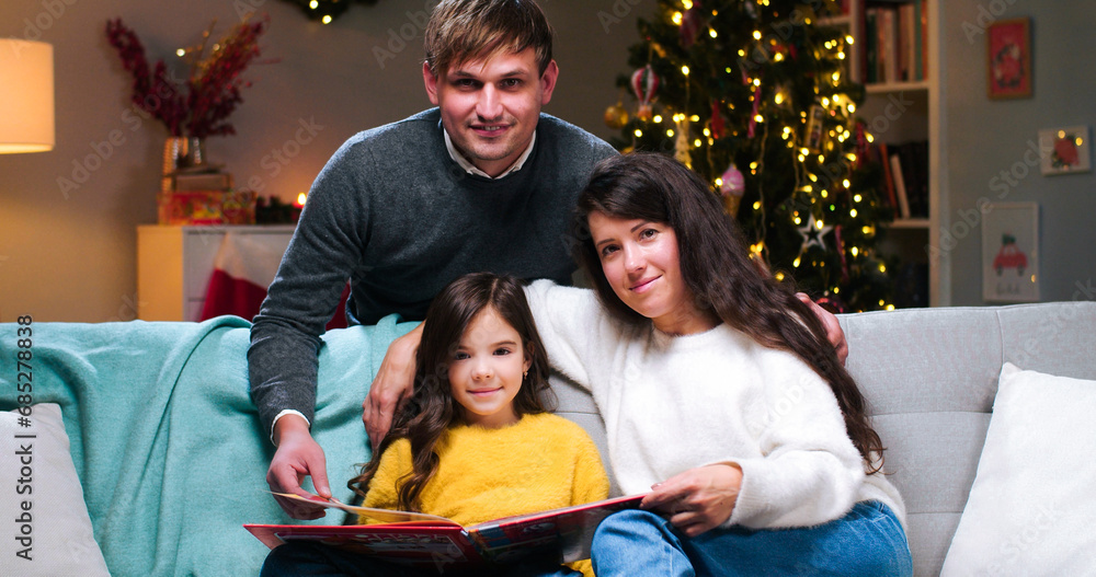 Portrait of lovely Caucasian cute little girl sitting at decorated room with happy parents reading book of fairy tales together. Mom and dad with kid on Christmas at home. New Year celebration concept