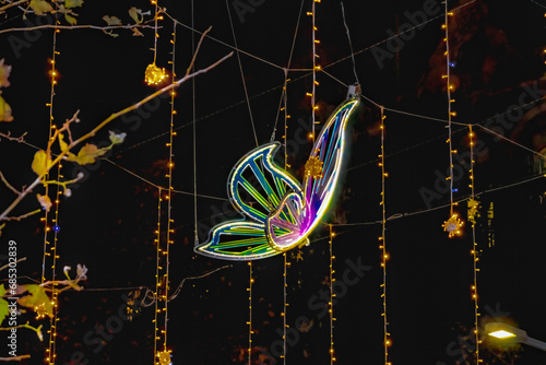 Photography Neon butterfly among Christmas garlands against the background of the black sky at dusk, close-up