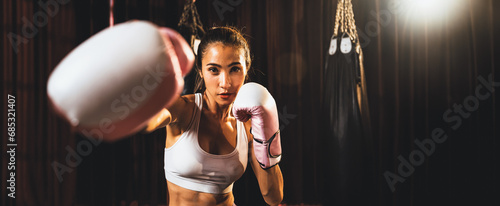 Asian female Muay Thai boxer punch fist in front of camera in ready to fight stance posing at gym with boxing equipment in background. Focused determination eyes and prepare for challenge. Spur