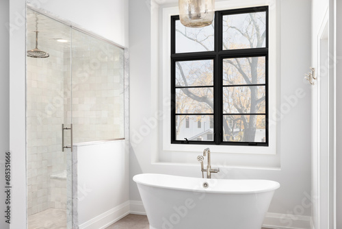 A bathroom detail with light hanging above a freestanding soaker tub  a black large framed window  and a tiled walk-in shower.