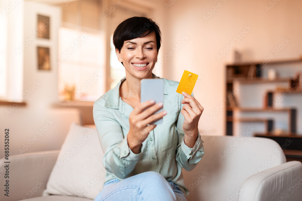 Cheerful middle aged lady holds smartphone and credit card indoors Cheerful middle aged lady holds smartphone and credit card indoors