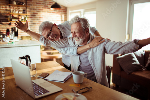 Happy senior man with outstretched arms looking at laptop with wife in home photo