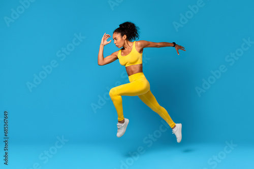 African Lady In Fitwear Exercising Jumping Over Blue Studio Background