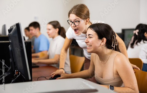 Two young girl student emotionally reacting looking at monitor while working at pc in computer class of college