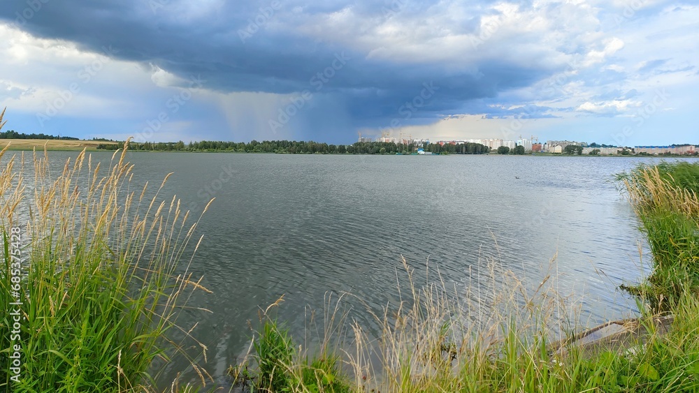 There is a storm cloud over the lake and it is raining. The water ripples in the wind. In summer, tall grass grows on the shore and reeds grow in the water. On the far shore are buildings and cranes