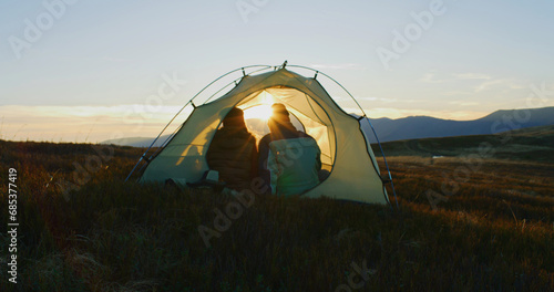 Multiethnic family sits in tent on top of hill and kisses. African American man clinks cups with his Caucasian wife and looks at sunset. Young tourists enjoy view on their vacation in mountains.
