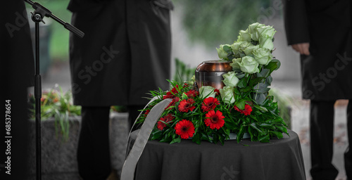 Metal urn or funeral container with ash of a deceased person at a memorial service. Undertakers seen in the back and a beautiful bouquet surrounding the urn. photo