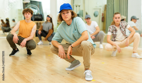 Modern confident teenager in blue cap posing during group training in hip hop studio, squatting on haunches with teammates on background. Selective focus