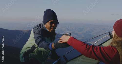 African American man sets up tent on top of mountain hill. Woman gives cup of tea to backpacker friend. Multiethnic family on adventure trip. Diverse tourist couple stopped to rest during a hike.