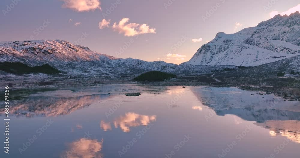 Aerial view from low flying drone of sea coast, snowy mountains in winter at sunrise. Lofoten islands, Norway. Top view of fjord, reflection, water, stones, snowy rocks, purple sky with clouds. Lake