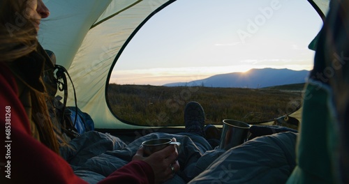 Close up of young Caucasian woman holding cup, talking with African American man and looking at sunset. Couple of tourists rest in tent on top of hill and enjoy view. Hikers in mountains on vacation.
