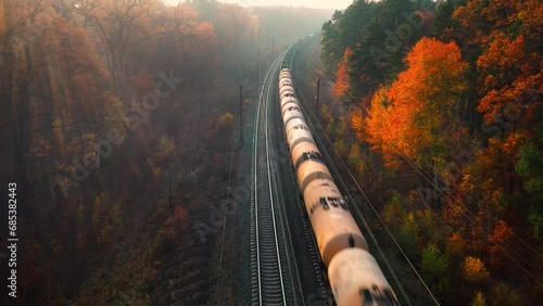 Aerial view of freight train in colorful forest in fog at sunrise in autumn. Top drone view of moving train with wagons in woods in fall. Industrial. Railroad, foggy trees with orange leaves. Railway