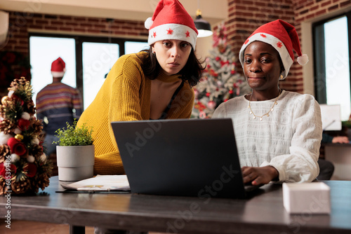 Company employee sitting at desk and coworking with colleague on project in christmas decorated corporate environment. Diverse women workers in xmas hats doing teamwork during winter holiday