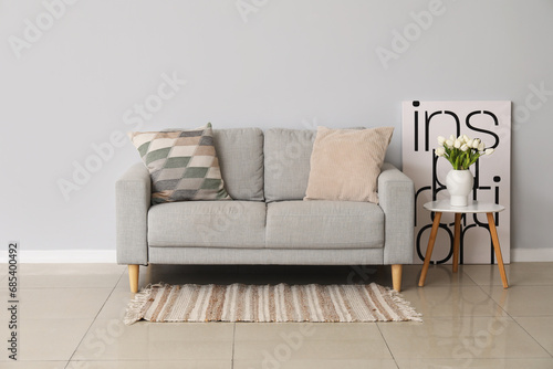 Cozy sofa with cushions and tulip flowers in vase on table near grey wall #685400492