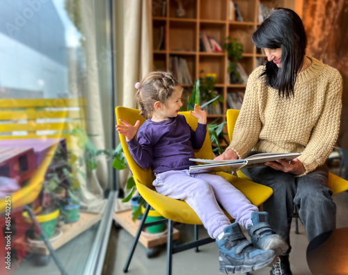 A grandmother reads a book to her granddaughter. A woman reads a book to a child, a girl, sitting in yellow chairs by the windo , family, joy, emotion photo