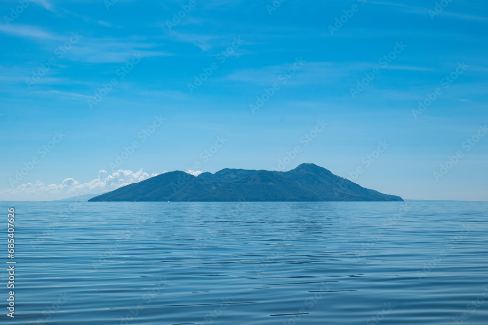 Island Seen From Afar with Volcanic Hills, Lush Vegetation Forest in Front of a Calm Ocean Island Seen From Afar with Volcanic Hills, Lush Vegetation Forest in Front of a Calm Ocean