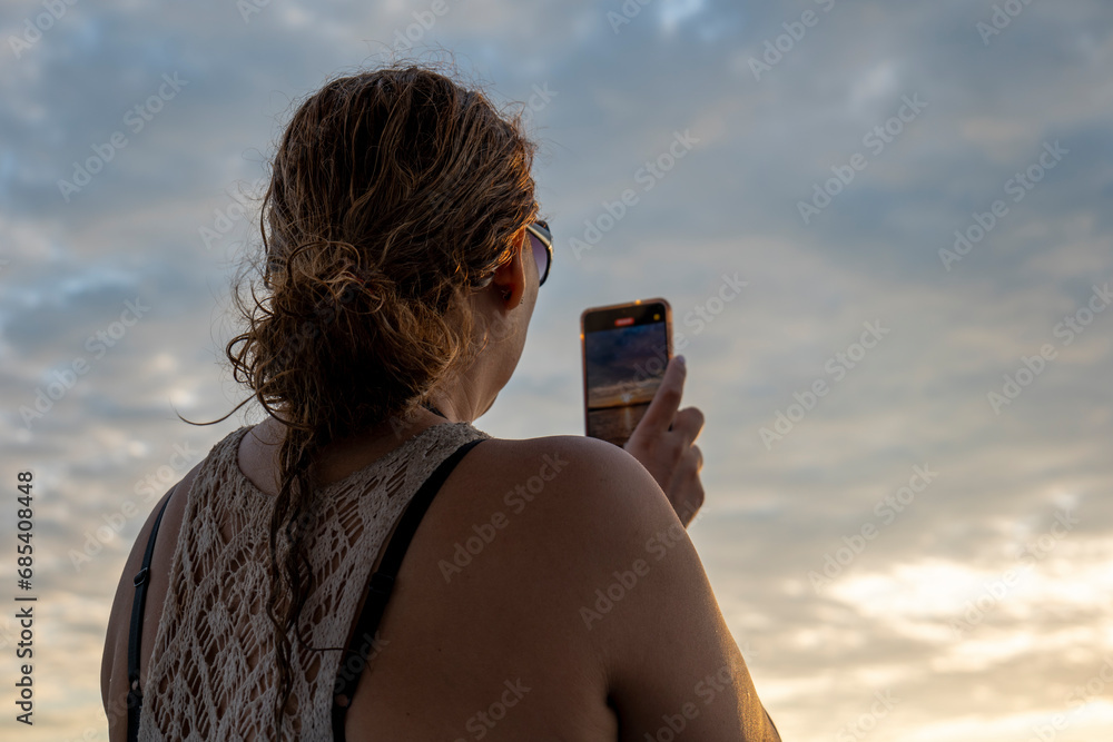 Adult Woman in a Sleeveless Shirt Holds a Smartphone to Take a Picture of the Sunset on a Boat Prior to Arrival in the Port