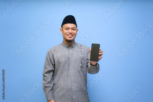 Asian Muslim man holding phone and pointing finger at mobile phone ready to pay zakat isolated on blue background. Islam lifestyle concept. Celebration Ramadan and eid Mubarak photo