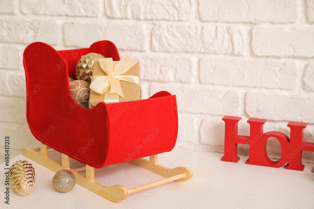 Obraz premium Decorative sleigh with Christmas decorations and gift box on table near light brick wall