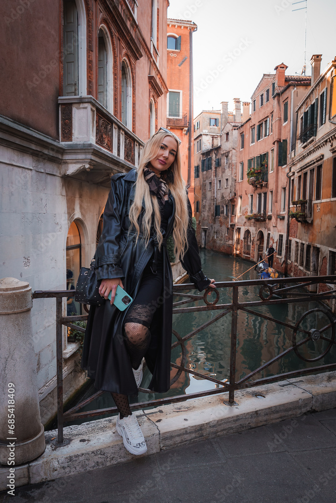A woman in a black leather jacket and white sneakers poses on a bridge in Venice, Italy, with gondolas and colorful buildings in the background, evoking a romantic and nostalgic ambiance.