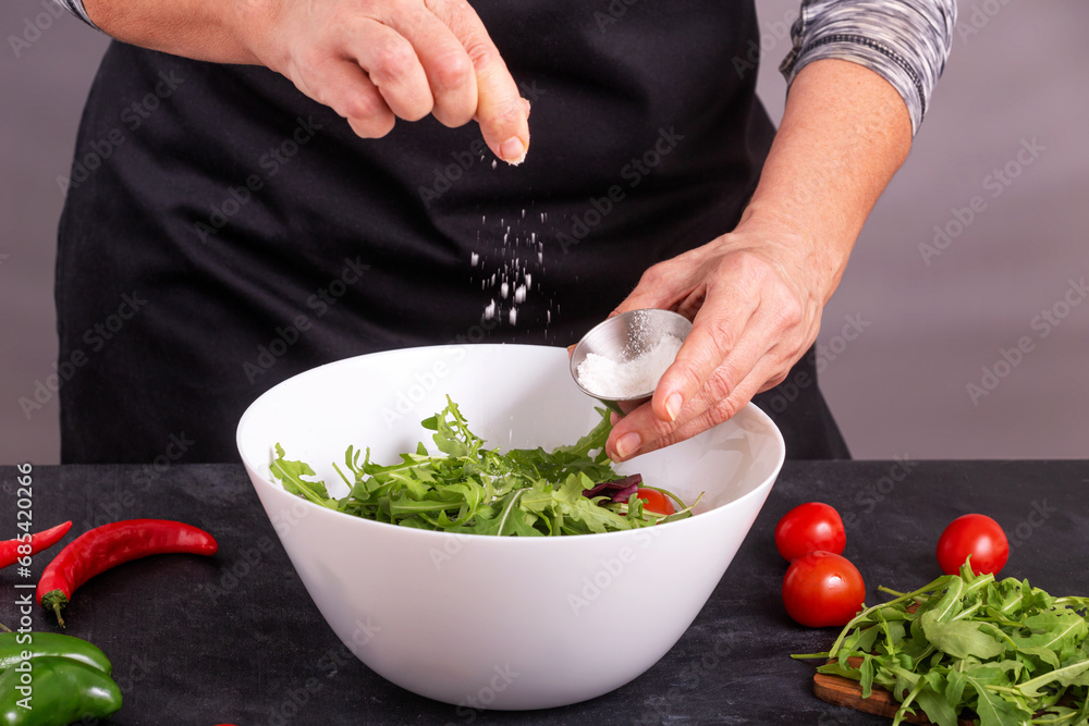 A cook sprinkles salt on a fresh vegetable salad with tomatoes and arugula, closeup with selective focus A cook sprinkles salt on a fresh vegetable salad with tomatoes and arugula, closeup with selective focus