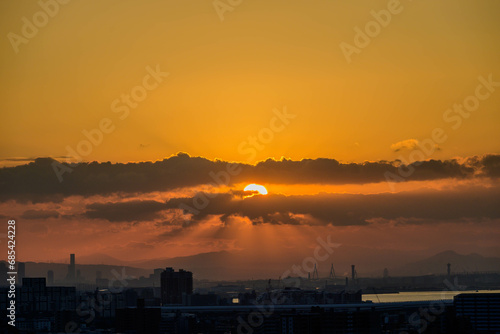 都市の夜明け。神戸市の高台から大阪湾と大阪南港から朝日が昇る。雲の隙間から天使の梯子。 © 宮岸孝守