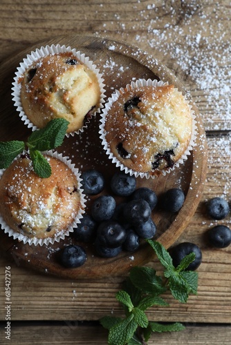 Delicious muffins powdered with sugar, blueberries and mint on wooden table, flat lay