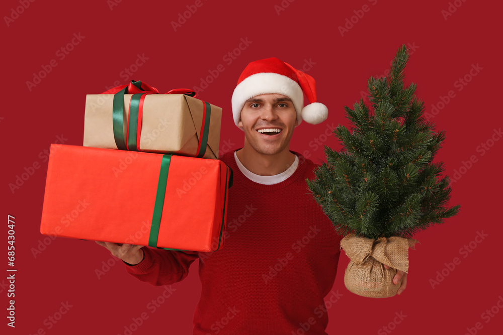 Young man in Santa hat with Christmas tree and gift boxes on red background Young man in Santa hat with Christmas tree and gift boxes on red background