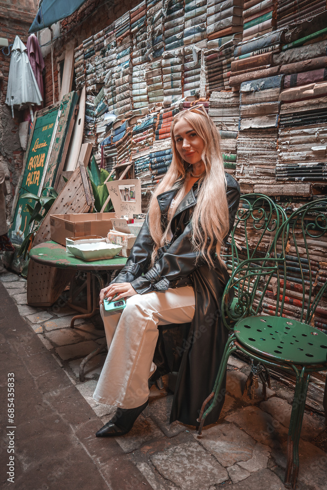 A woman sits in front of a wall of books in an unspecified location, possibly Venice, Italy. The image captures a serene moment surrounded by literature.