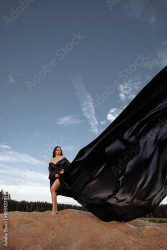 A brunette in a black dress with a long train made of silk fabric stands on the sand in the desert in the movement of the wind. Landscape of desert and blue sky with clouds. 
