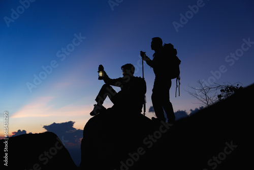 Silhouette of Asian two male standing raised hands with trekking poles and kerosene black lamp on cliff edge on top of rock mountain with at sunset rays over the clouds background,