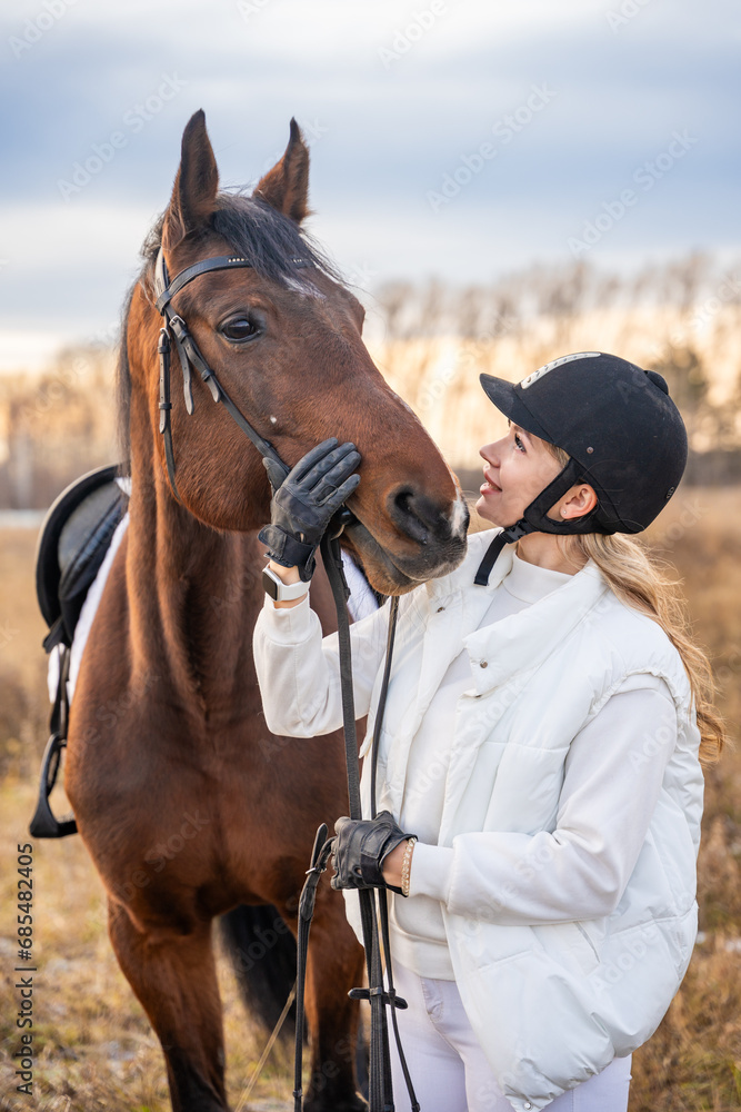 Fototapeta premium Beautiful blond professional female jockey standing near horse in field. Friendship with horse concept
